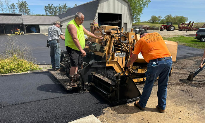 New-Asphalt-2 Davis Construction workers lay asphalt with a paving machine in a rural setting.